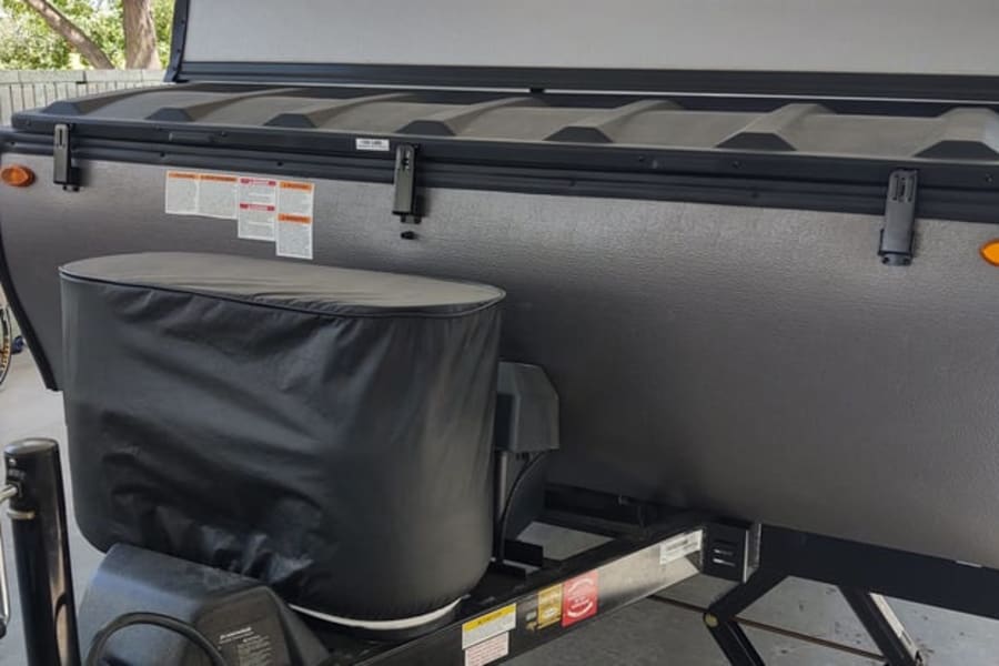 This RV rental's front view: a covered propane tank on the hitch, with warning labels on the gray, textured pop-up camper body.