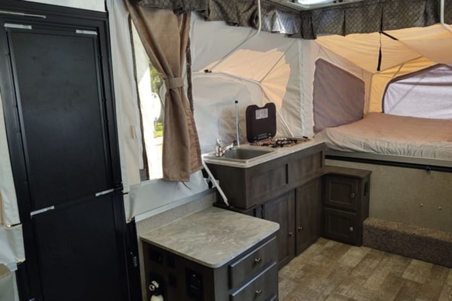 Interior of a pop-up rv rental, showing the kitchenette with a sink & stove, a bed, and the entrance door.