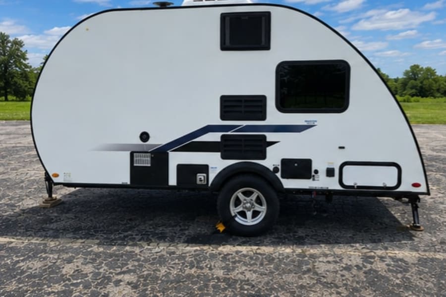 This white teardrop-style **RV rental** offers compact travel. Parked on asphalt with trees & blue sky in the background.