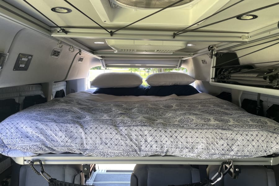 Inside an RV rental: a cozy bed with patterned bedding, overhead storage system, and fan for comfortable van life travel.