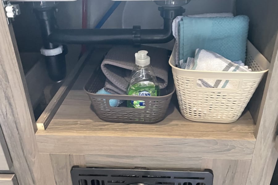 Organized under-sink storage in an RV rental, with dish soap, towels in baskets, plumbing, and a floor vent.
