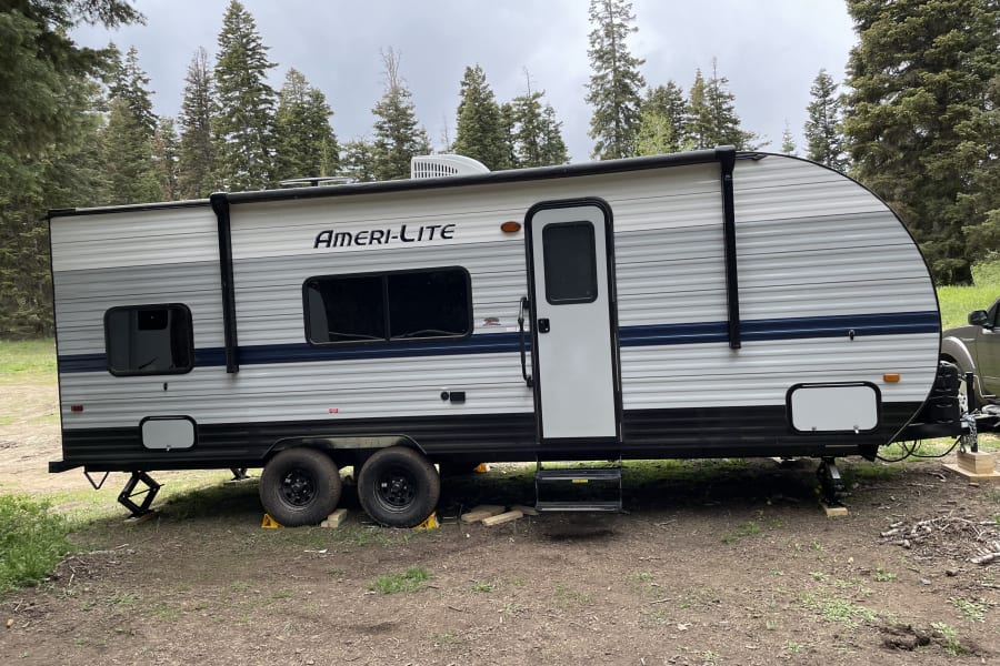 Ameri-Lite travel trailer (RV rental) parked in a forest campsite, set up for outdoor adventures among tall pine trees.