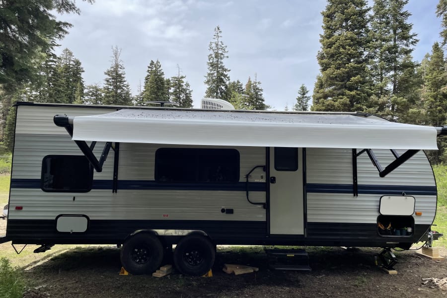 RV rental travel trailer parked at a wooded campsite, awning extended for a relaxing outdoor experience.