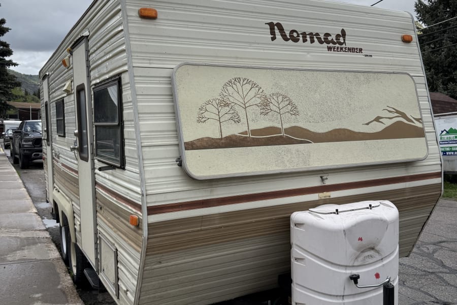 This Nomad Weekender vintage travel trailer, an ideal rv rental, is parked roadside under a cloudy sky.