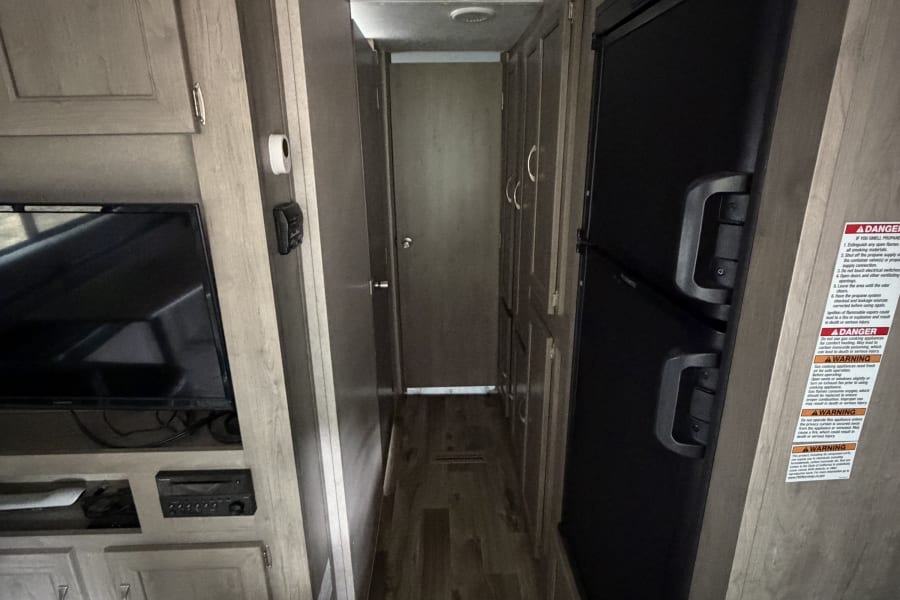 View of an RV rental interior hallway. A refrigerator, TV, and cabinets line the passage leading to a closed door.