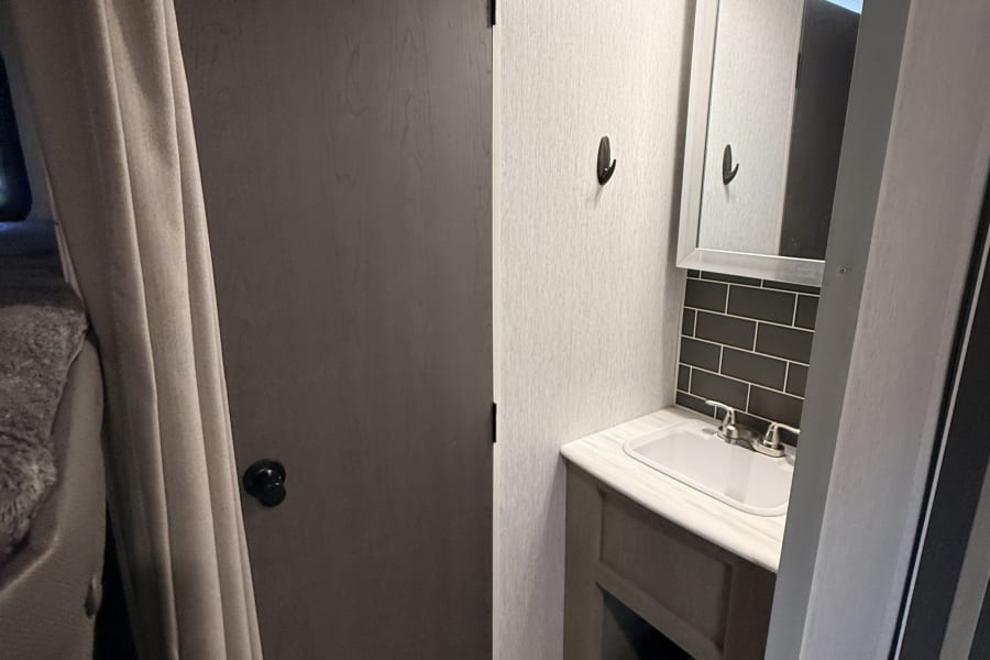 RV rental bathroom with sink, mirror, and dark tiled backsplash, next to a bedroom privacy curtain.