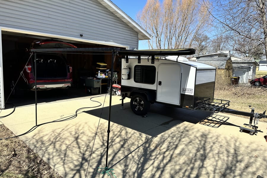 A compact Hiker camping trailer, perfect for an rv rental, set up with an awning outside a home garage on a sunny day.