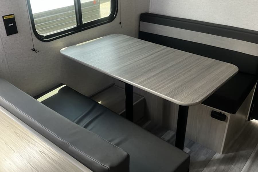 Modern RV rental dinette with light wood table & dark bench seating, by a window. Ideal for comfortable travel trailer living.