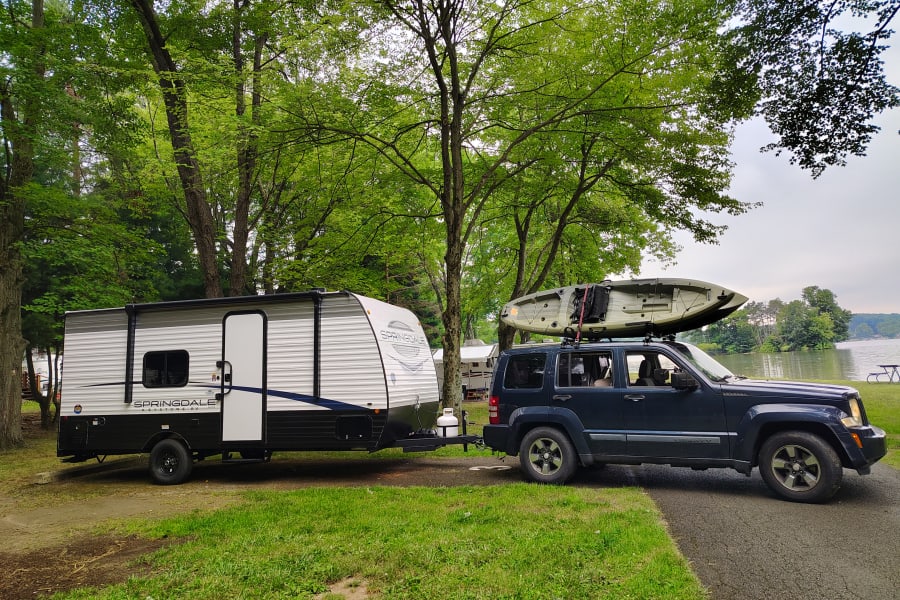 An RV rental with a kayak-laden SUV at a scenic lakeside campsite under lush trees, ready for outdoor adventures.