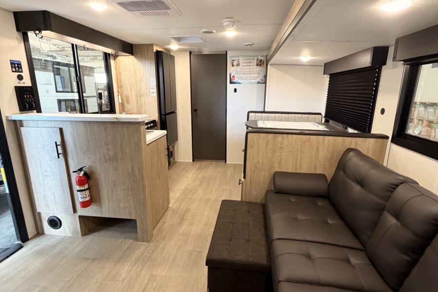 Modern rv rental interior with an L-shaped sofa, kitchenette, and dinette, featuring light wood-look flooring.