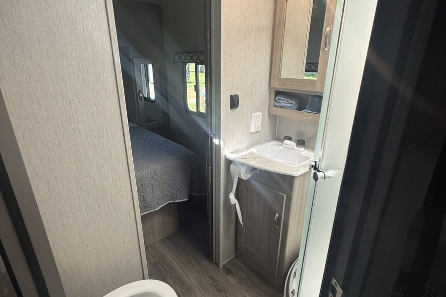 Inside a bright RV rental: compact bathroom with toilet, sink, and shower, offering a glimpse into the adjoining bedroom.