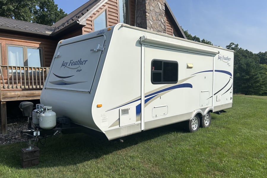 This white Jayco Feather EXP travel trailer is an ideal rv rental, parked on green grass next to a log cabin home.
