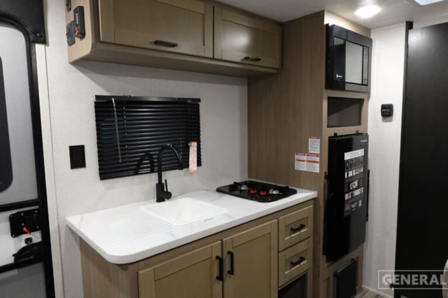 Inside an RV rental: a compact kitchen with sink, two-burner stove, microwave, refrigerator, and storage cabinets.