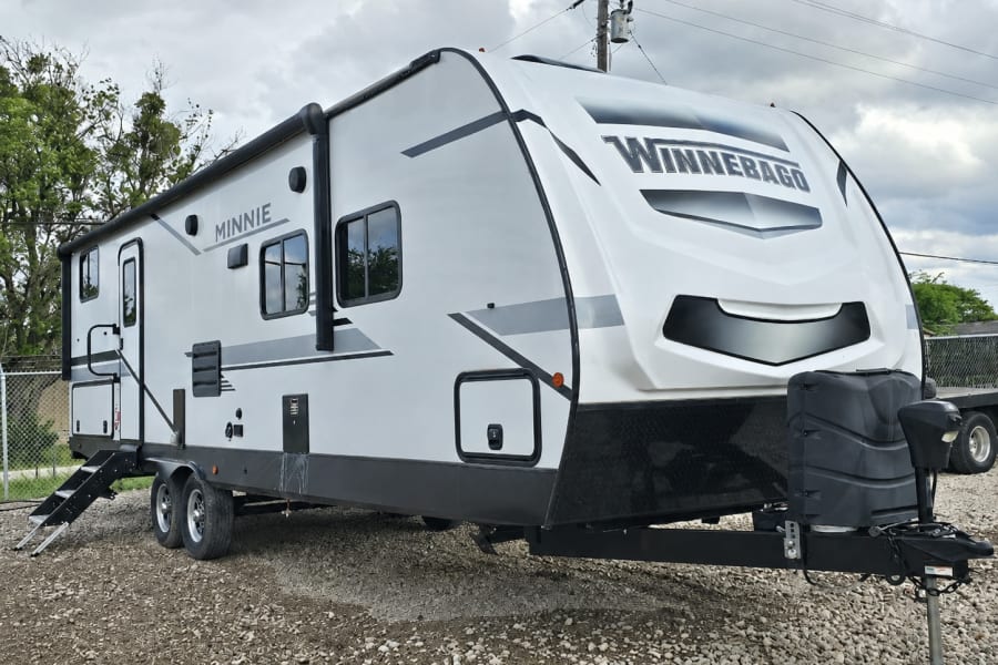 Get ready for adventure with this white Winnebago Minnie travel trailer rv rental, parked on gravel with steps down.