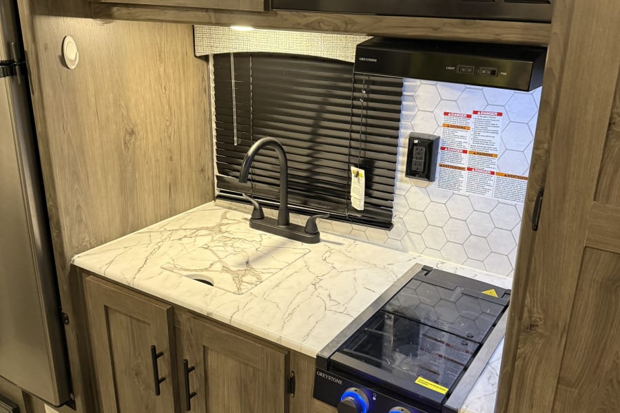 Bright RV rental kitchen features a microwave, sink, and stovetop. Modern wood-grain cabinets and marble-look countertops.