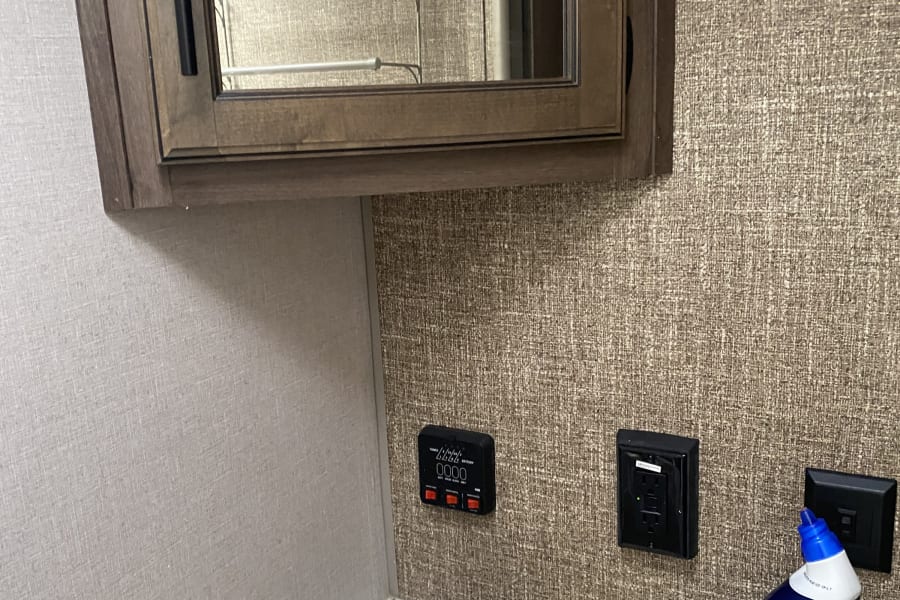 Inside an RV rental bathroom, featuring a vanity with sink, mirror, and a multi-gauge tank/battery monitor. Compact and practical.