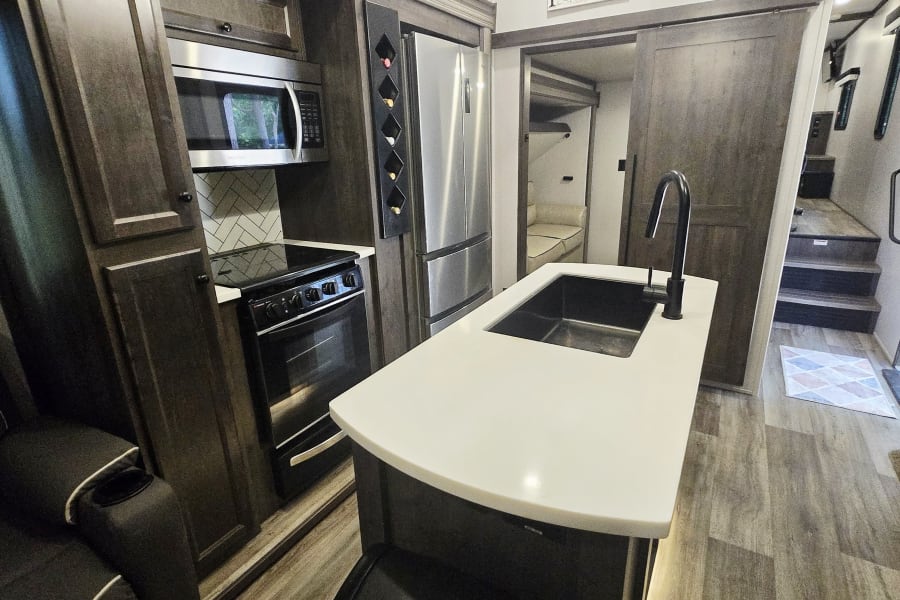 Modern RV rental kitchen: dark wood cabinets, stainless appliances, white island with sink. Bunkhouse and pantry access visible.