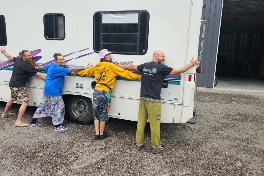 Playful group effort: four people push a white RV rental on a gravel lot, showing teamwork.