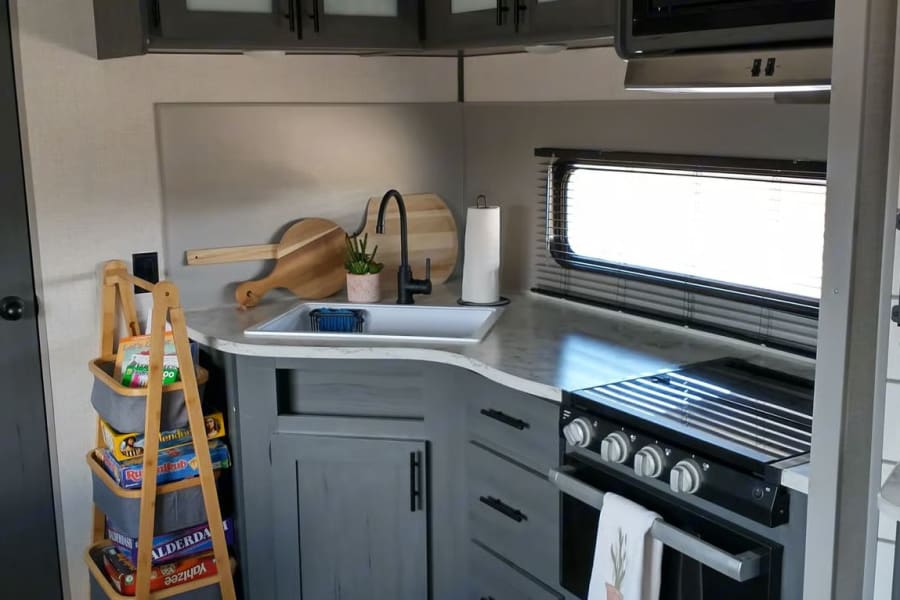 Inside an rv rental, a modern kitchen with gray cabinets, sink, stove, window, and games in a storage rack.