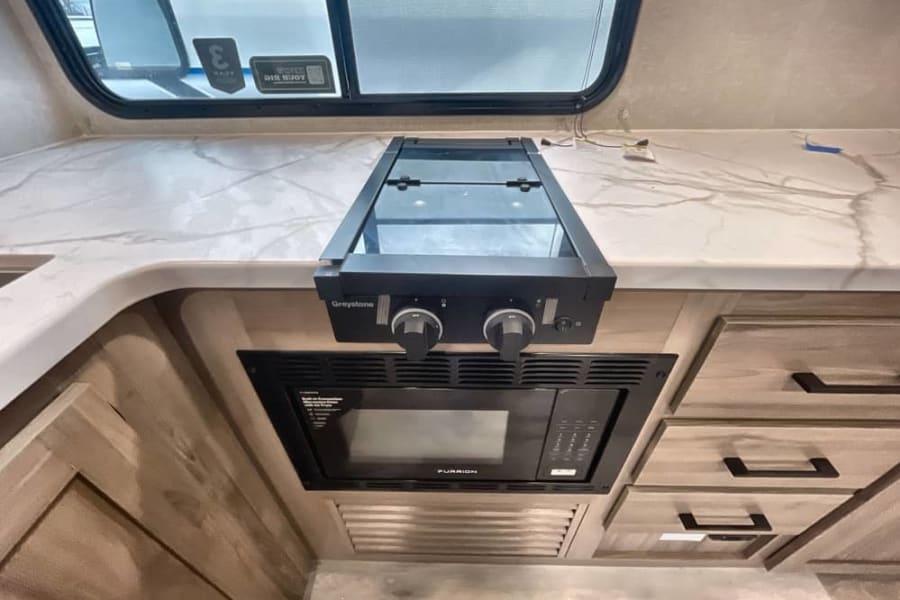 Inside an RV rental: a sleek kitchen featuring a white marble-look countertop, built-in Greystone cooktop, and Furrion microwave.