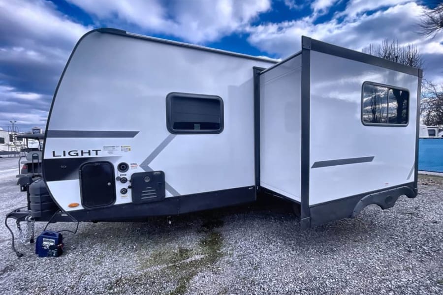 A bright white travel trailer with a side slide-out extended, ready for your next rv rental adventure. Parked on gravel.