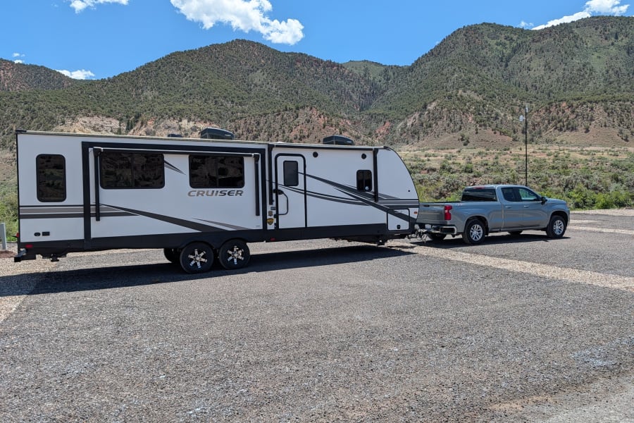 This white RV rental and gray pickup truck are ready for adventure, parked in a scenic mountain setting under a blue sky.