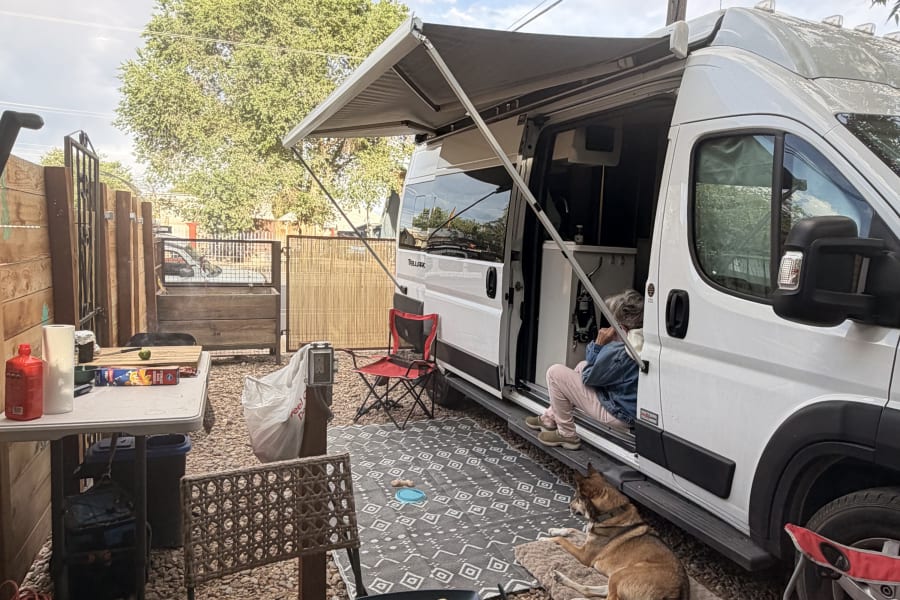Relaxing RV rental evening: A person inside, a dog resting outside, with snacks and drinks on a mosaic table under an umbrella.