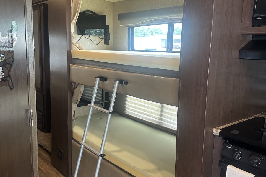 An RV rental interior shows cozy bunk beds with a ladder, window, and TV, adjacent to a compact kitchen stove.