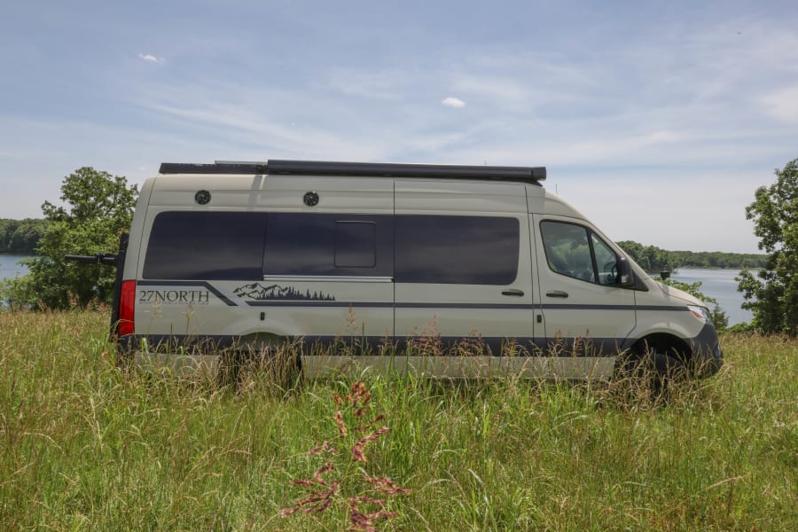 A light-colored RV rental camper van, branded "27NORTH", sits in tall grass beside a lake, ideal for outdoor adventures.