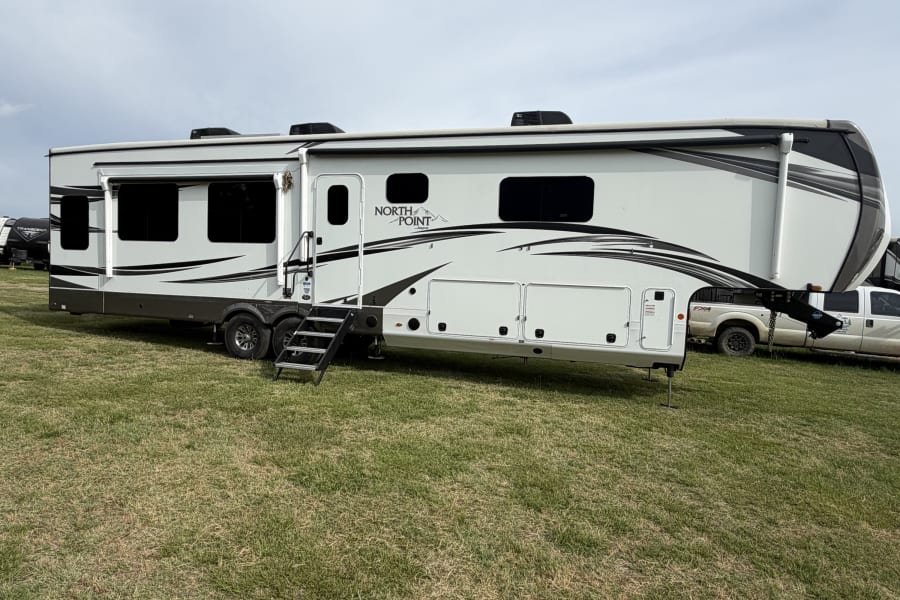 A large white and gray North Point fifth-wheel rv rental with steps extended is parked on green grass under a cloudy sky.