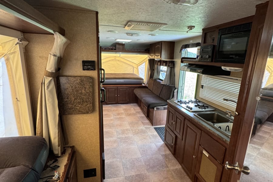 RV rental interior: kitchen with stove/sink, sofa, and pop-out tent bed. Features tiled floor & wood cabinets, compact design.