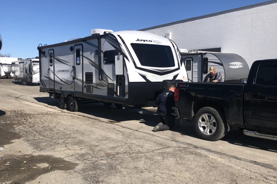 A man preps a Jayco RV rental, working on the hitch of the travel trailer connected to a black truck in an outdoor RV lot.