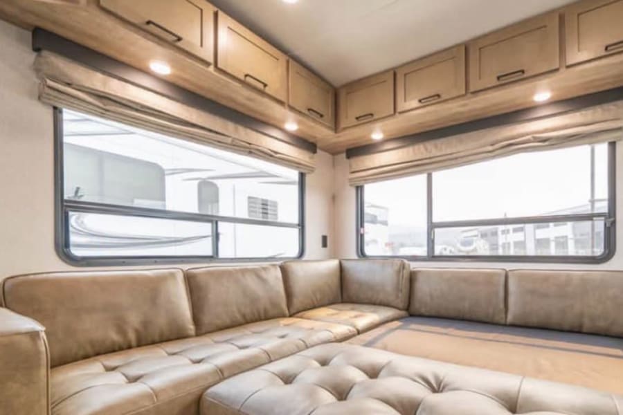 Cozy RV rental living space with a large, convertible sectional sofa, two bright windows, and wood cabinets.