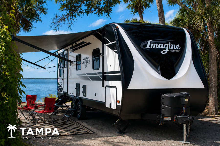 Enjoy this 'Imagine' travel trailer RV rental by the water. Featuring an awning, lights, and red chairs for your perfect camping trip!