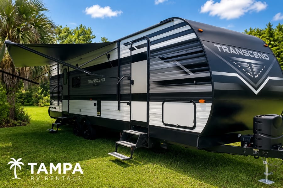 Spacious Transcend travel trailer rv rental under a blue sky, perfect for adventure. Extended awning on green grass.
