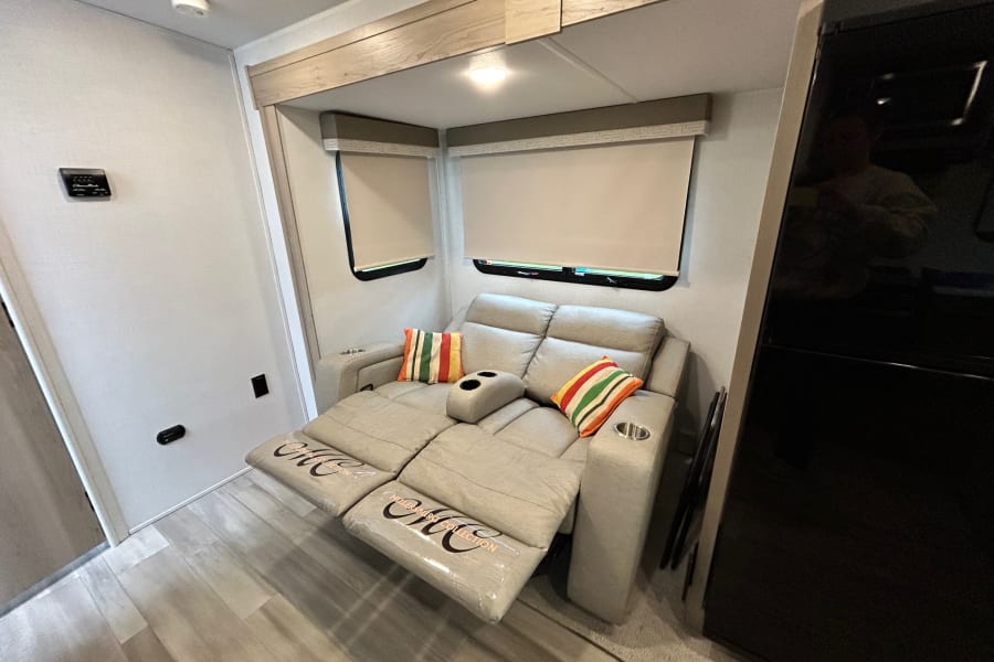 Cozy RV rental interior featuring a modern reclining sofa with cup holders, colorful pillows, and roller blinds for comfort.