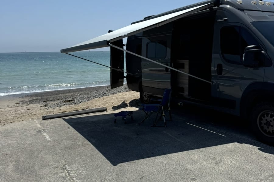 Enjoy a sunny beach day with an rv rental! A gray camper van is parked with its awning extended by the ocean.