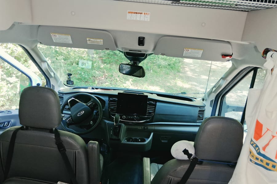 Inside an RV rental: driver's view of the dashboard, steering wheel, and green trees outside. Overhead storage.