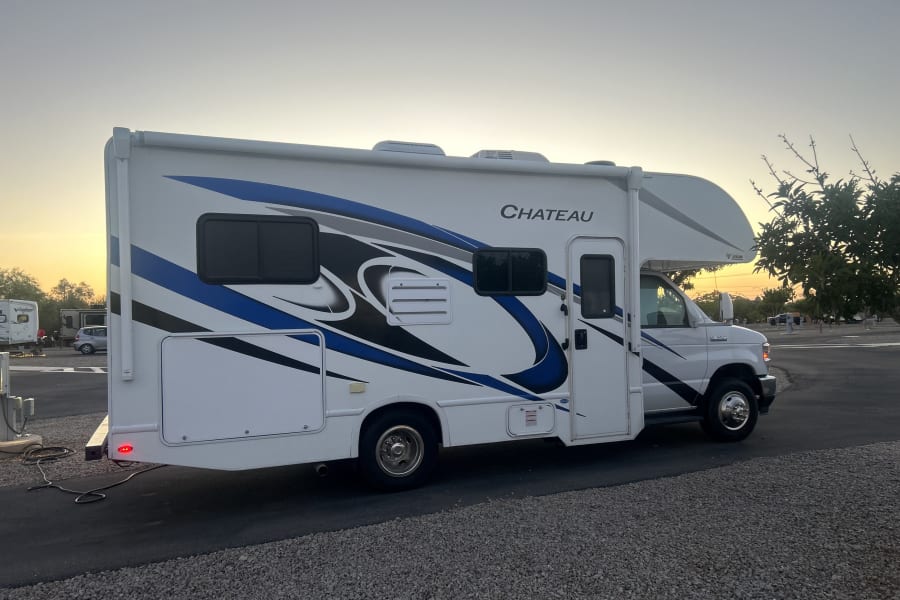 A Chateau RV rental, white with blue and black graphics, parked on a gravel site at dusk. Power cord connected for camping travel.