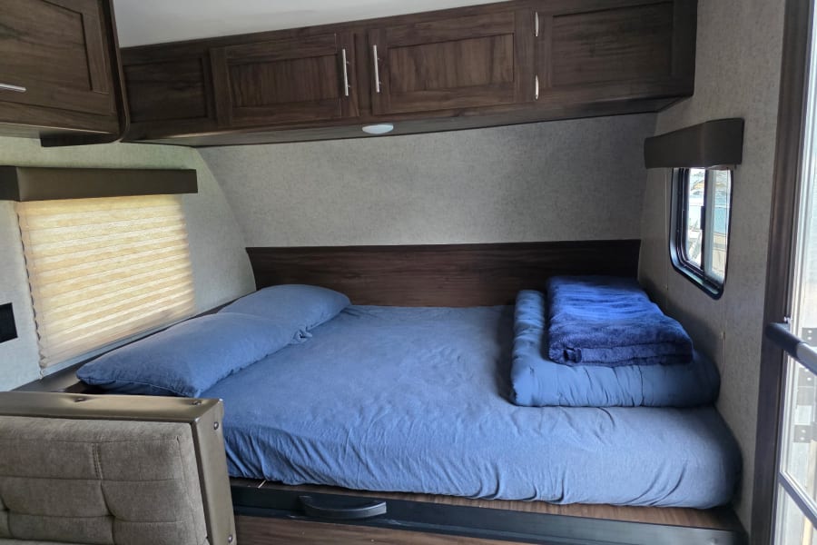 Inside this RV rental, a comfy sleeping area features blue bedding, overhead cabinets, and a window for natural light.