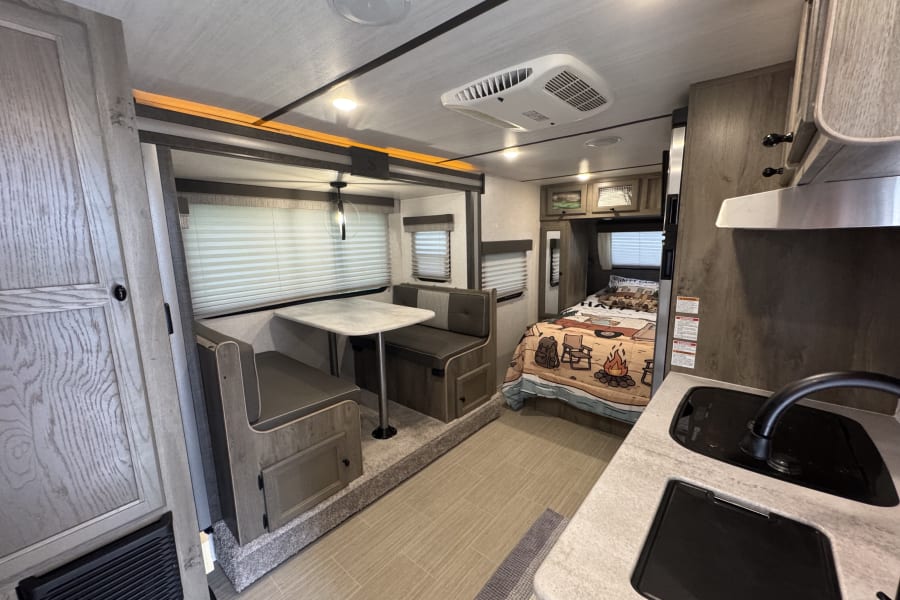 RV rental interior: dinette, compact kitchen with sink/stove, and a cozy bedroom with camping-themed bedding. Modern, functional design.