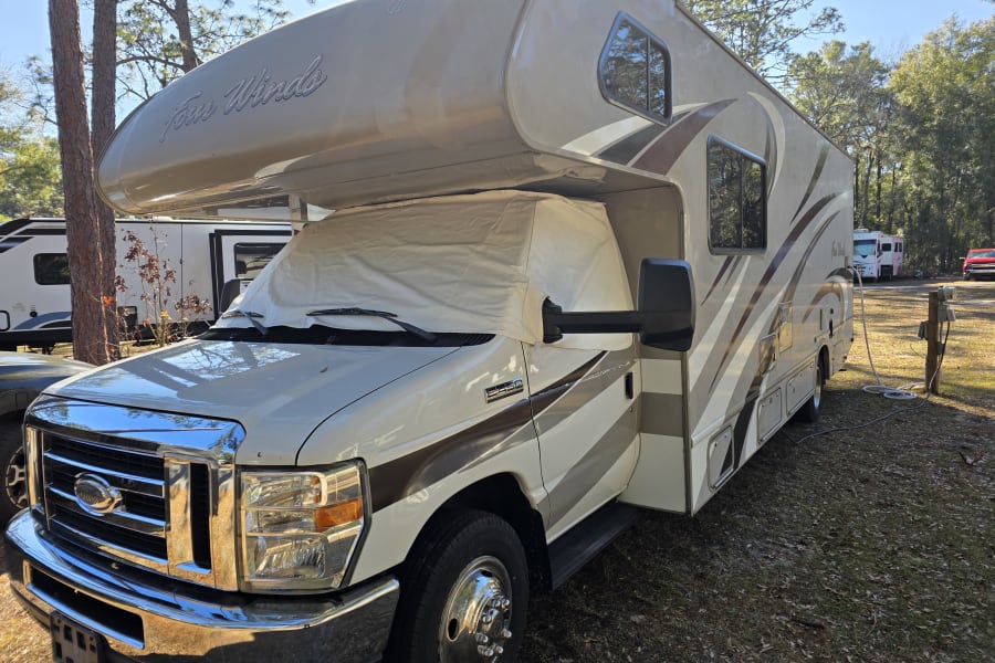 Class C Four Winds rv rental parked in a sunny campground, connected to utility hookups, with its windshield covered.