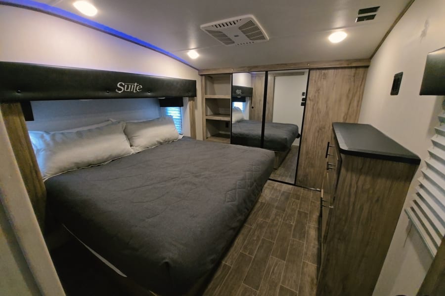 This rv rental bedroom features a "Suite" headboard, large bed, mirrored closet, wooden dresser, and blue LED ceiling lights.