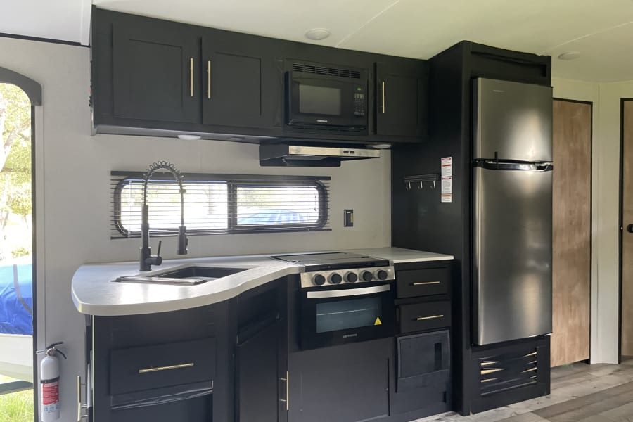 This modern rv rental kitchen boasts dark cabinets, a stainless fridge, microwave, stove, and sink. Compact & functional.