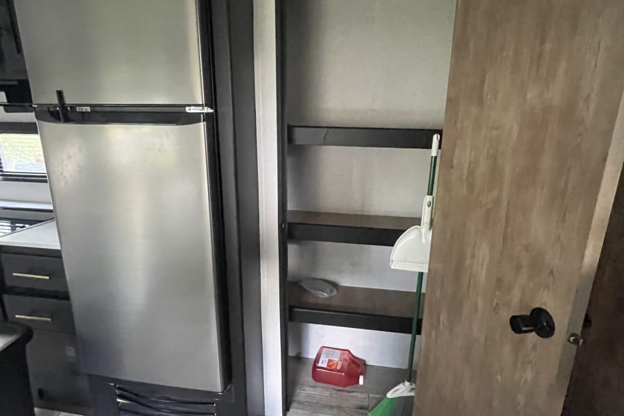 Inside an RV rental: a compact stainless steel fridge, shelved pantry with cleaning supplies, and wood-look floor.