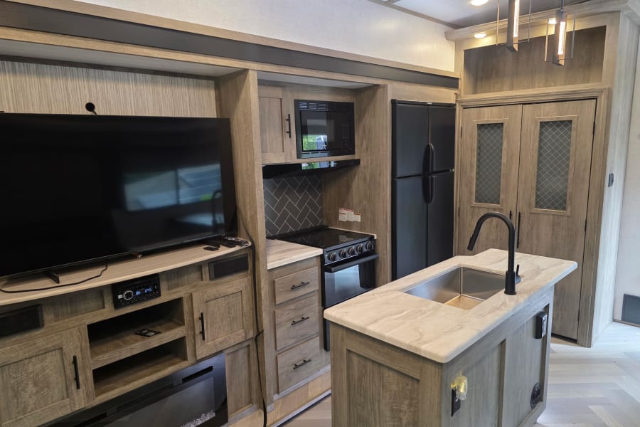 Modern RV rental interior with a light wood kitchen featuring an island, black appliances, a TV, and electric fireplace.