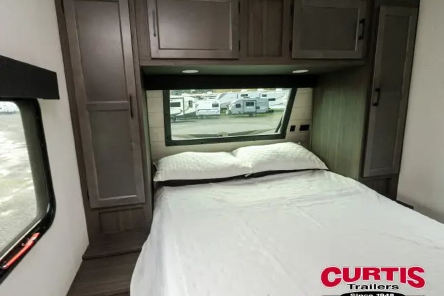 RV rental bedroom with bed, overhead cabinets, and a window view of other RVs, offering cozy accommodation.