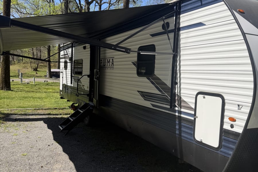 A Puma travel trailer RV rental with its awning extended, parked on a gravel campsite amidst trees. Ideal for outdoor adventures.