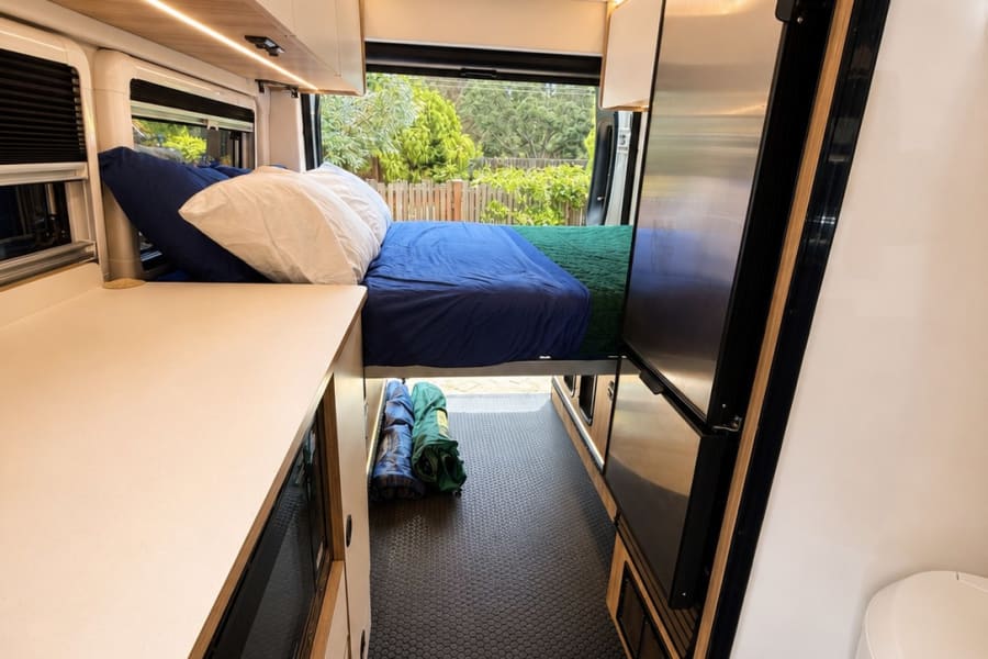 RV rental: Modern camper van interior with a cozy bed, sleek kitchen counter, large fridge, and compact bathroom with a toilet.