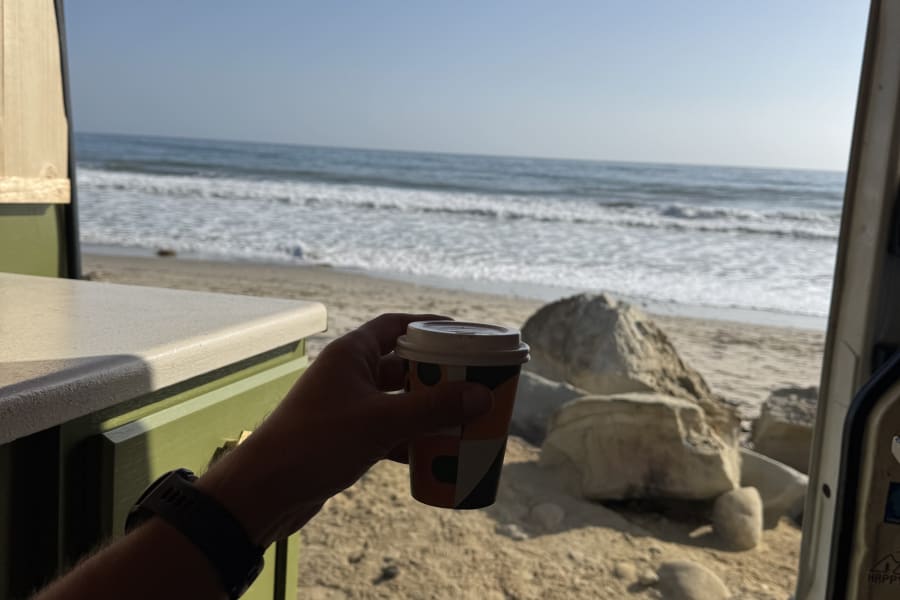 Sipping coffee from an RV rental, a person overlooks a sunny beach with gentle waves and scattered rocks.